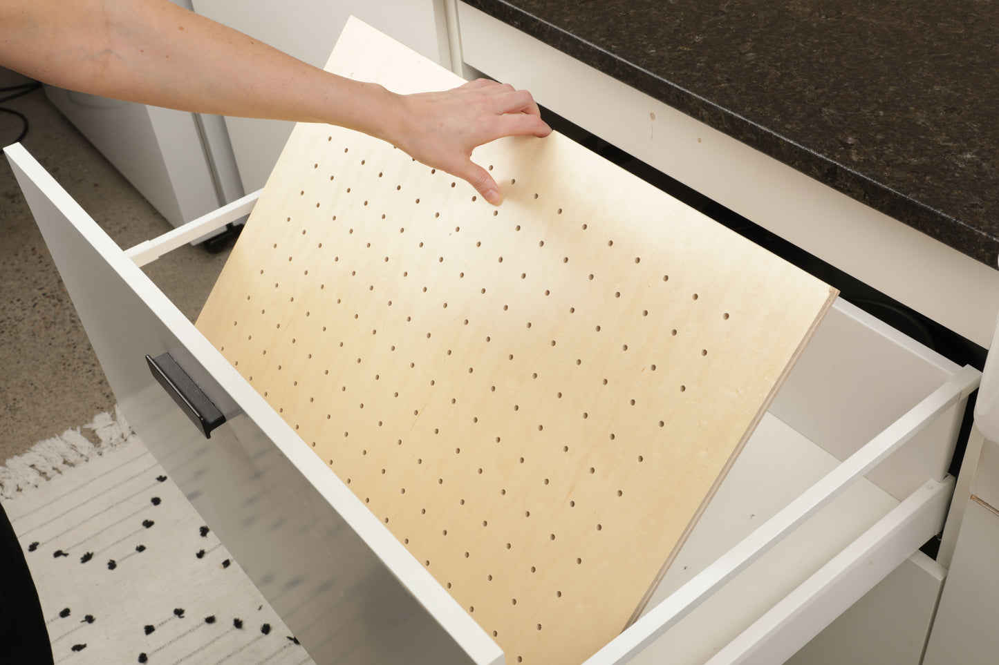 A person's hand is reaching into a white drawer to pull out a wooden board with holes, which appears to be a storage solution for organizing items.