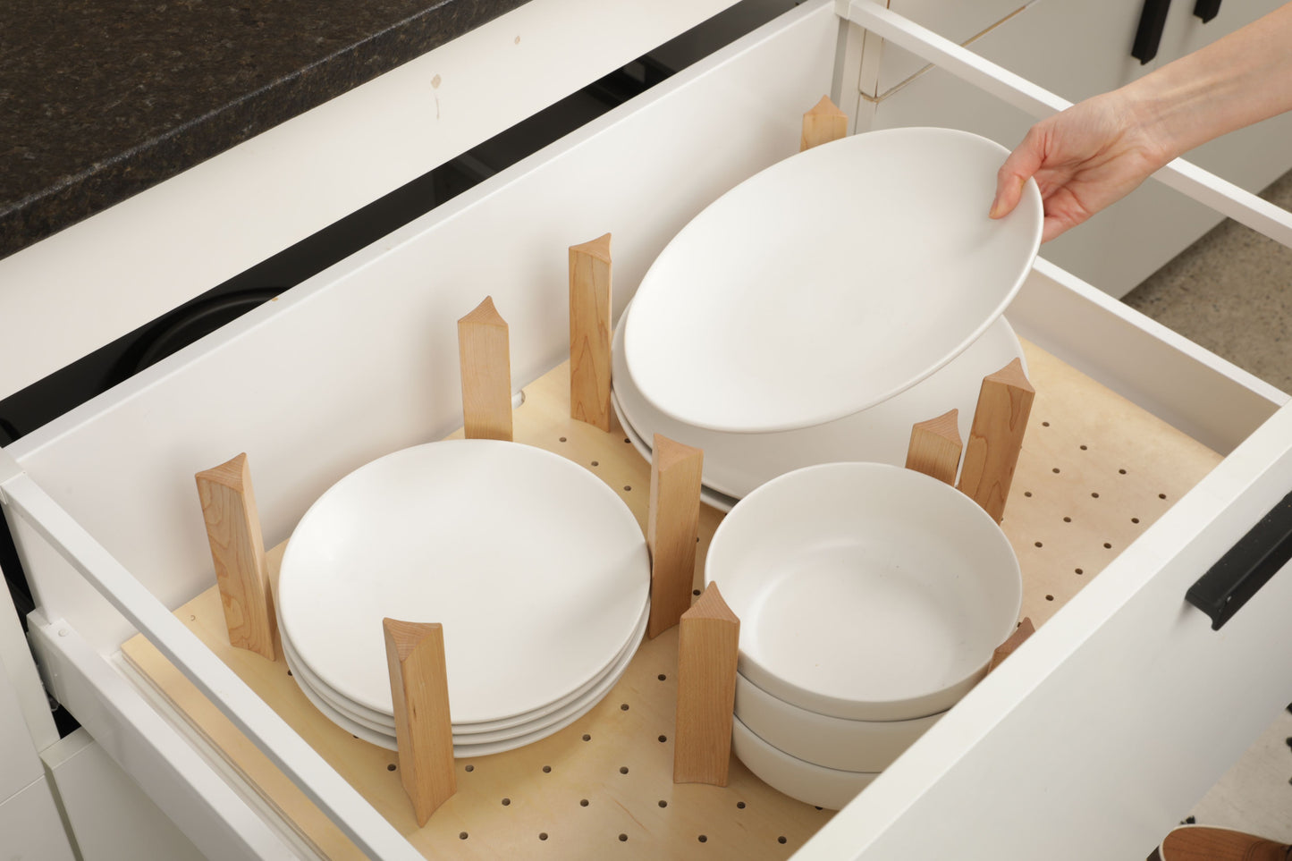 A person's hand is reaching for a white plate from a drawer with wooden dividers, which are neatly arranged inside the drawer.
