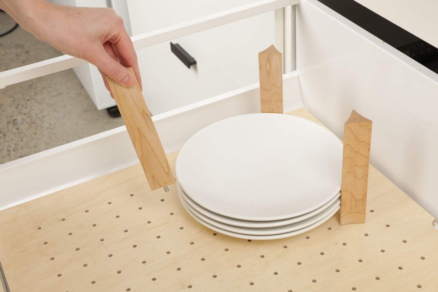 A hand is placing wooden blocks on a wooden shelf, which is holding a stack of white plates.